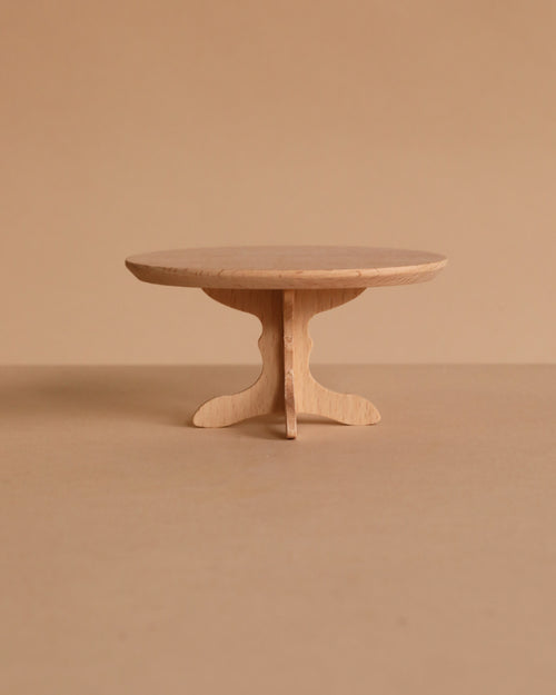 The PRE-ORDER: Maileg Oval Dining Table, Mouse Size - Nature features a small, round wooden design with a single pedestal base, displayed on a plain beige surface that highlights SuitCasa’s signature wood quality.