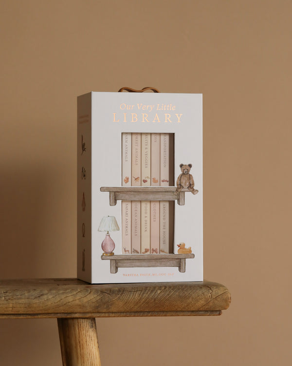 The "Our Very Little Library Board Book Set" sits on a wooden stool, featuring shelves filled with vocabulary-building baby board books, accompanied by a teddy bear, lamp, and small figurines. The scene is set against a warm beige background.