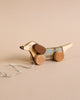 A Handmade Pull Along Dog Toy shaped like a dachshund with a checkered pattern on its body, round wheels painted with natural milk paint, and a white string against a beige background.