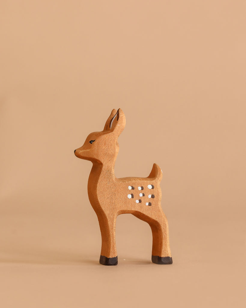 The Bumbu Handmade Wooden Fawn, featuring delicate white spots, stands on a beige surface against a matching beige background, facing left.