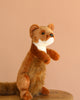 The Weasel Stuffed Animal, a plush toy with realistic brown weasel features, sits upright on a wooden surface against a plain beige background.