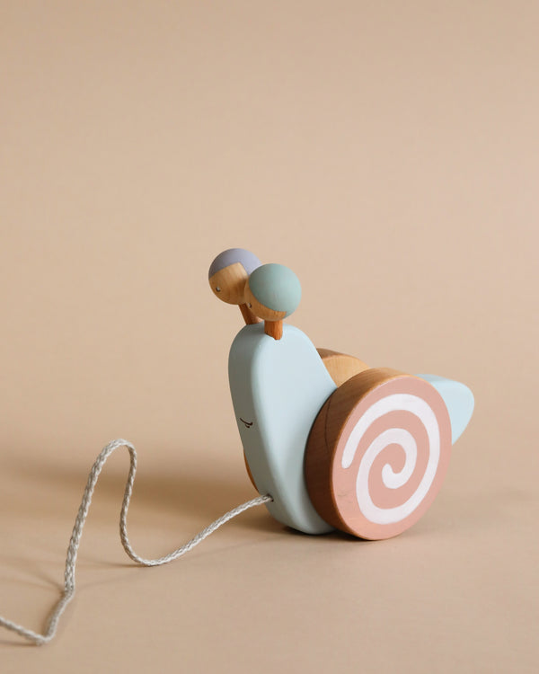 A Handmade Pull Along Snail Toy crafted from natural materials, with a light blue body and natural wood accents, featuring a pink and white shell, on a beige background.