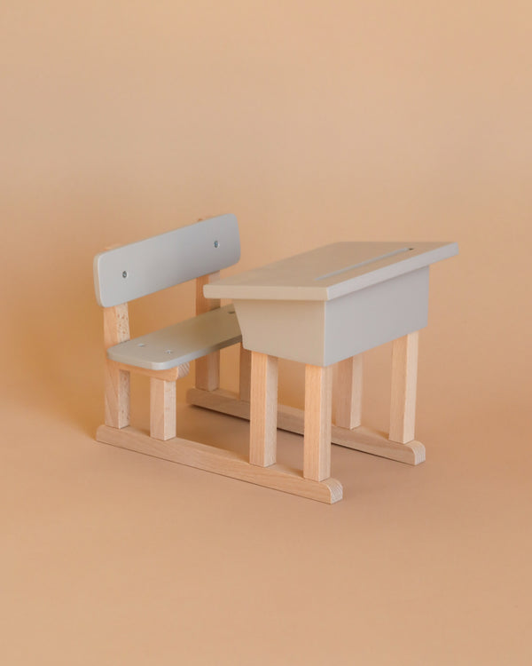 The Minikane Doll School Desk – Grey is a small, minimalist model featuring both a wooden school desk and bench set. Expertly crafted by an artisan carpenter, each piece showcases light wood and grey accents. The desk includes a slanted surface with an indented slot for holding pencils, all set against a plain peach background. Ideal for displaying Minikane Babies.