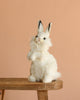 The White Bunny Stuffed Animal sits upright on a wooden stool against a peach background, highlighting its realistic plush design and quality craftsmanship.