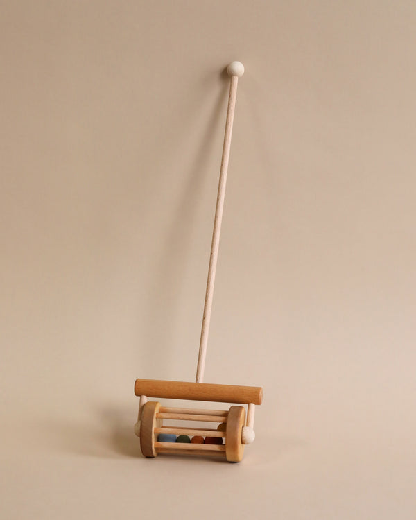 A Handmade Push Toy Rattle standing upright against a beige background. The instrument consists of a long stick made from sustainably harvested birch wood attached to a wheeled base that contains several loose wooden cylinders.