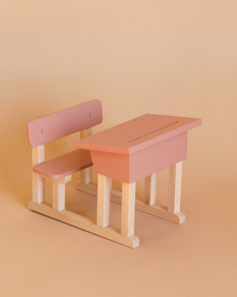 The Minikane Doll School Desk in Powder Pink, paired with a matching bench, is photographed against a plain beige background. The desk features a slanted top while the bench includes a simple backrest, creating an aesthetic that is both minimalist and playful—an ideal addition for children or as an accessory for Minikane Babies.