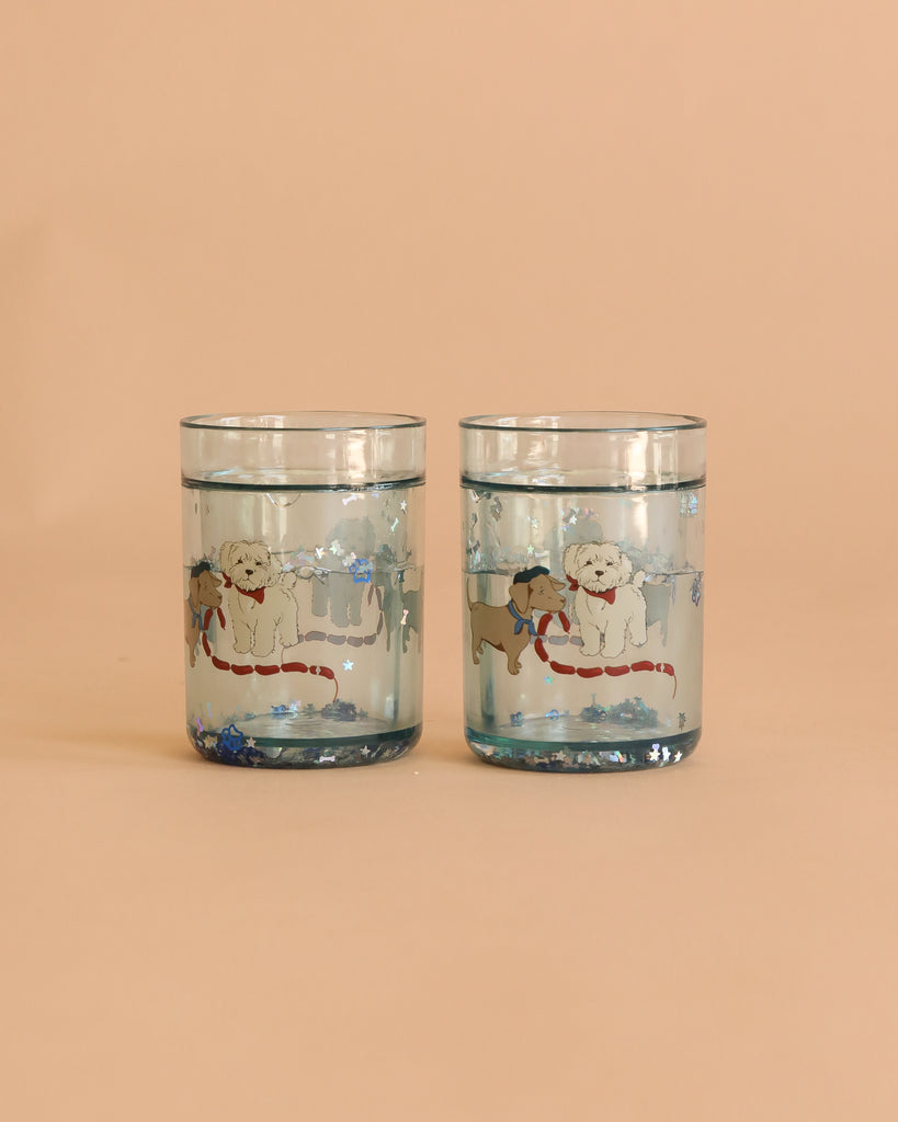 The Konges Sloejd Glitter Cups - Today's Special (2 Pack) features clear cups with a white and a brown dog in scarves on snowy backgrounds. Blue glitter accents create magical kids' drinkware perfect for adding sparkle to every sip.