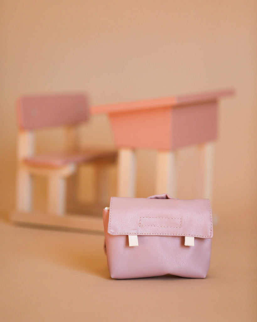 A pink Minikane Doll School Bag made of faux leather from the Back to School Collection is placed on a beige surface, with an out-of-focus pink and beige toy desk and chair set in the background. The image, featuring soft pastels, perfectly complements Minikane Gordis dolls.