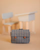 A Minikane School Bag - Flannel, a small plaid bag with a handle and labeled "MADE IN FRANCE," sits on a light brown surface. Behind it, a blurred miniature wooden school desk and bench painted white enhance the scene. The background is also light brown, creating a soft, minimalist aesthetic perfect for Minikane Gordis dolls.
