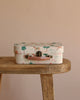 A closed Maileg On The Go Suitcase Set, featuring a small pastel-colored design with charming animal and tree illustrations, rests on a wooden bench against a beige background.