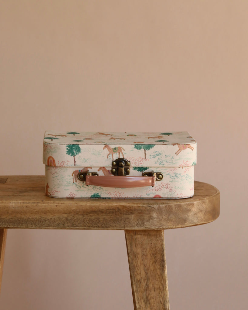A closed Maileg On The Go Suitcase Set, featuring a small pastel-colored design with charming animal and tree illustrations, rests on a wooden bench against a beige background.