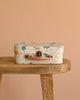 The Maileg Little House Living Room Travel Set, a pastel suitcase with animal and tree prints, rests on a wooden bench next to Maileg mouse accessories, all set against a plain beige background.