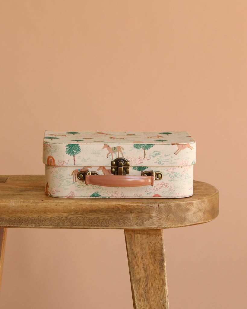 The Maileg Little House Living Room Travel Set, a pastel suitcase with animal and tree prints, rests on a wooden bench next to Maileg mouse accessories, all set against a plain beige background.