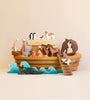 A collection of Ostheimer Handmade Large Wooden Ark, including giraffes, bears, and penguins, artistically arranged on and around a wooden boat against a peach background.