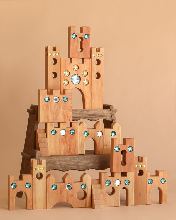 The Bauspiel Large Fairytale Castle (16 pieces) features wooden blocks with blue and gold gem accents, arches, turrets, and keyhole windows, creating a magical setting for imaginative play against a simple beige backdrop.