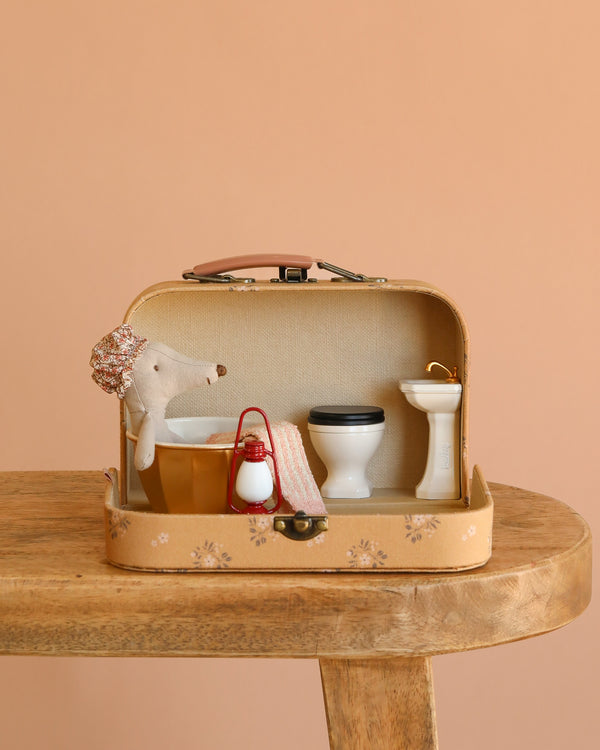 The Maileg Little House Bathroom Set, displayed in a small suitcase on a wooden table, includes miniature mouse furniture—a toilet, sink, brown tub, red lantern, and a mouse plush toy in a shower cap against a light peach background.