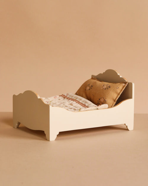 The Maileg Wooden Bed, Mouse - Off White features a gold floral pillow and a patterned blanket, shown against a beige background.