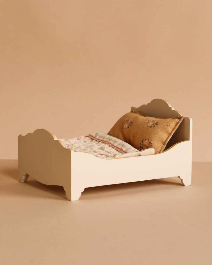 The Maileg Bedroom Set features a small white wooden bed with a beige patterned blanket and two gold decorative pillows, displayed against a plain beige background.
