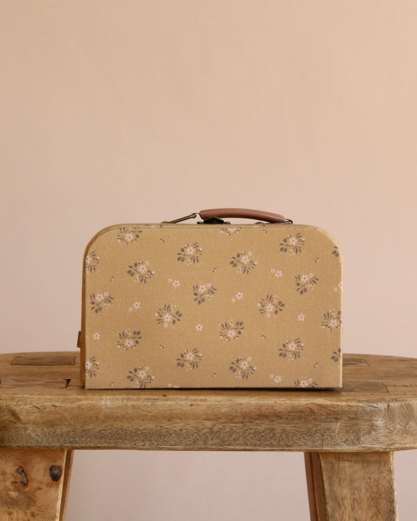 The Maileg Little Sister Mini House Set with a beige floral suitcase and brown handle rests on a rustic wooden table against a plain beige background.