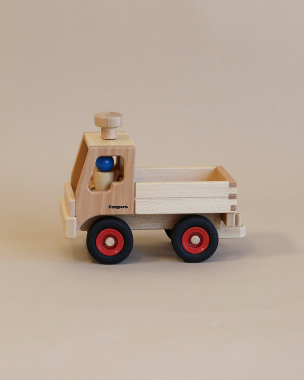 A handcrafted Fagus Wooden Unimog Truck with red wheels and a blue peg figure driver inside, set against a plain beige background.