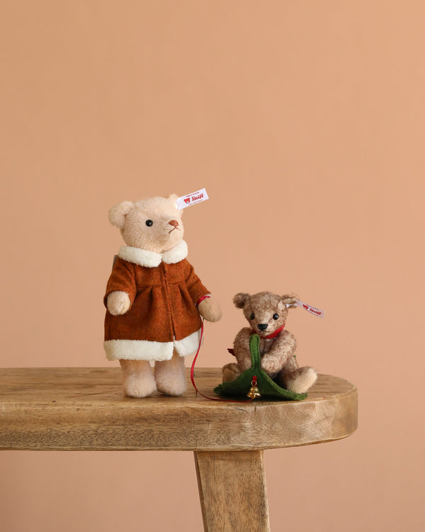 Two Steiff Mama Teddy Bear With Child collectibles sit on a wooden bench. Mama Bear wears a brown coat, the child holds a green Christmas tree, and both have ear tags, set against a solid light brown background.