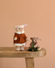 Two Steiff Mama Teddy Bear With Child collectibles sit on a wooden bench. Mama Bear wears a brown coat, the child holds a green Christmas tree, and both have ear tags, set against a solid light brown background.