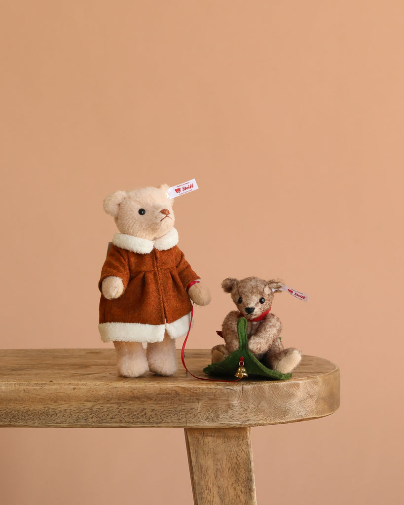 Two Steiff Mama Teddy Bear With Child collectibles sit on a wooden bench. Mama Bear wears a brown coat, the child holds a green Christmas tree, and both have ear tags, set against a solid light brown background.