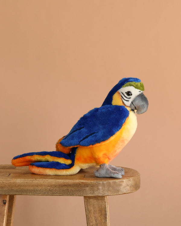 The Steiff Lori Parrot, a plush stuffed animal with vibrant blue, yellow, and orange feathers, sits on a wooden stool against a plain light brown background.
