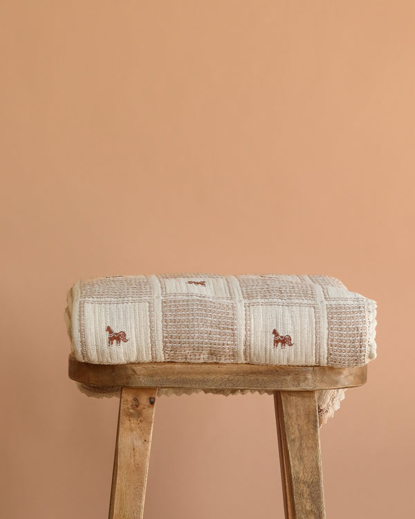 The Organic Cotton Twin Size Blanket - Pony, a folded beige knit with small brown pony designs, rests on a rustic wooden stool against a plain light brown background.