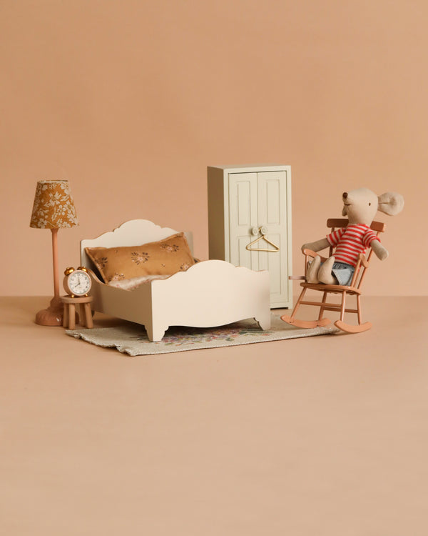 The Maileg Bedroom Set includes a miniature mouse on a rocking chair, a white bed with brown pillow, lamp, clock, white wardrobe, and a toy mouse in a red striped shirt—all on a beige background.