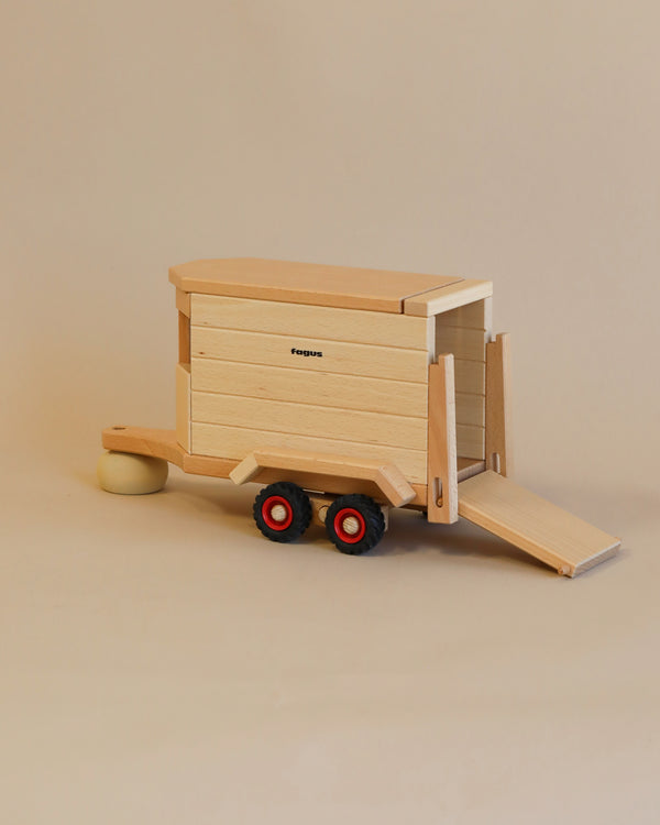 A Fagus wooden horse box with a ramp down, displaying red wheels and a hitch, set against a plain beige background.
