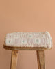 The Organic Cotton Twin Size Blanket - Fleur, featuring a light pink floral design, is folded on a wooden stool against a peach background. Made in Portugal from organic cotton.
