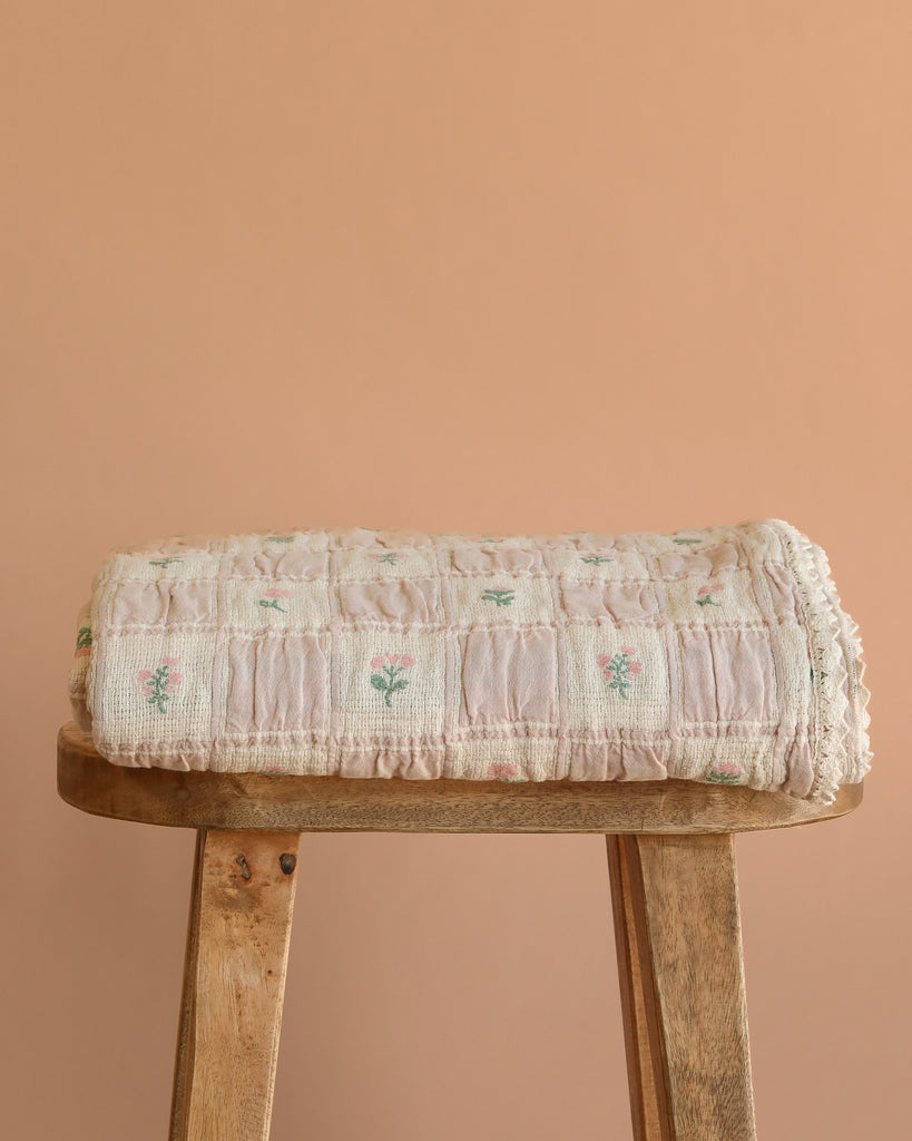 The Organic Cotton Twin Size Blanket - Fleur, featuring a light pink floral design, is folded on a wooden stool against a peach background. Made in Portugal from organic cotton.
