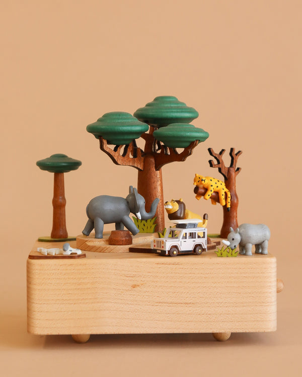 The Wooden Safari Music Box displays miniature safari animals—a cheetah in a tree, elephant, rhino, hippo, and a safari jeep—set among green trees on a beige background.