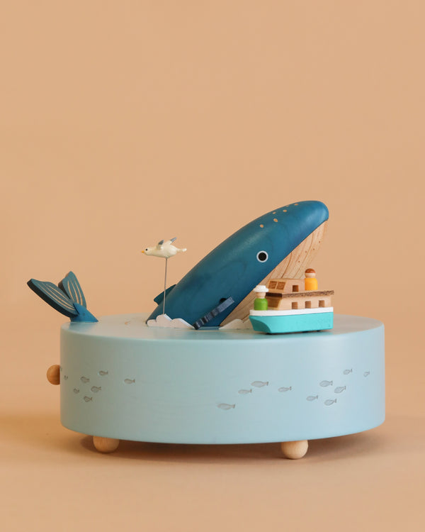 The Wooden Whale Music Box is a blue, ocean-themed wooden box with a whale, seagull, and small boat on top. Its round light blue base features tiny fish painted over a beige background.