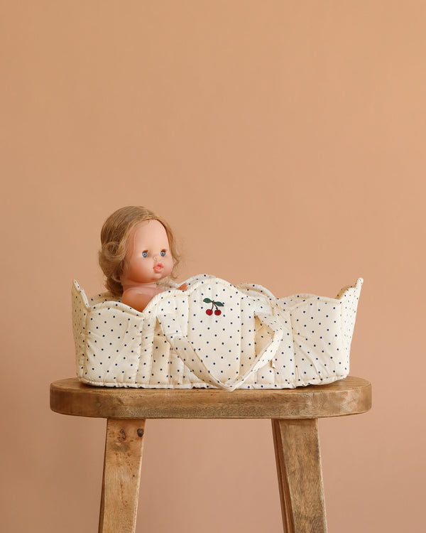 The Konges Sloejd Doll Lift - Estate Dot, featuring a cream polka-dot OEKO-TEX cotton design with cherries, holds a blonde-haired doll and sits on a wooden stool against a beige background.