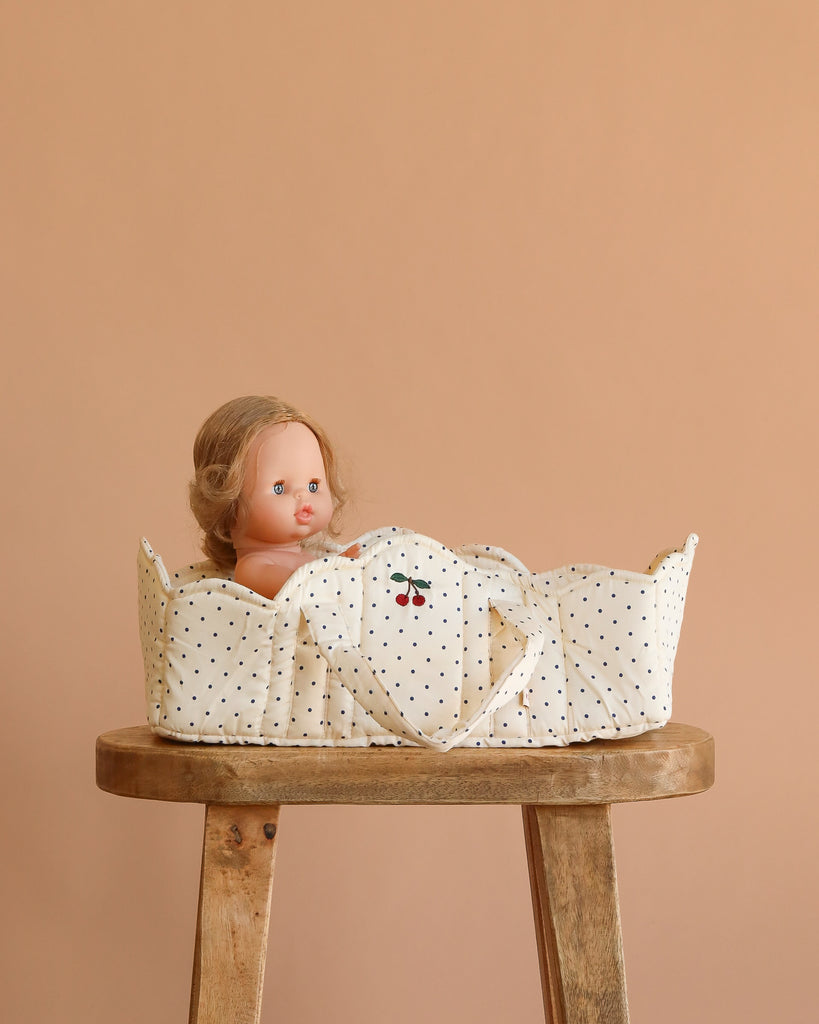 The Konges Sloejd Doll Lift - Estate Dot, featuring a cream polka-dot OEKO-TEX cotton design with cherries, holds a blonde-haired doll and sits on a wooden stool against a beige background.