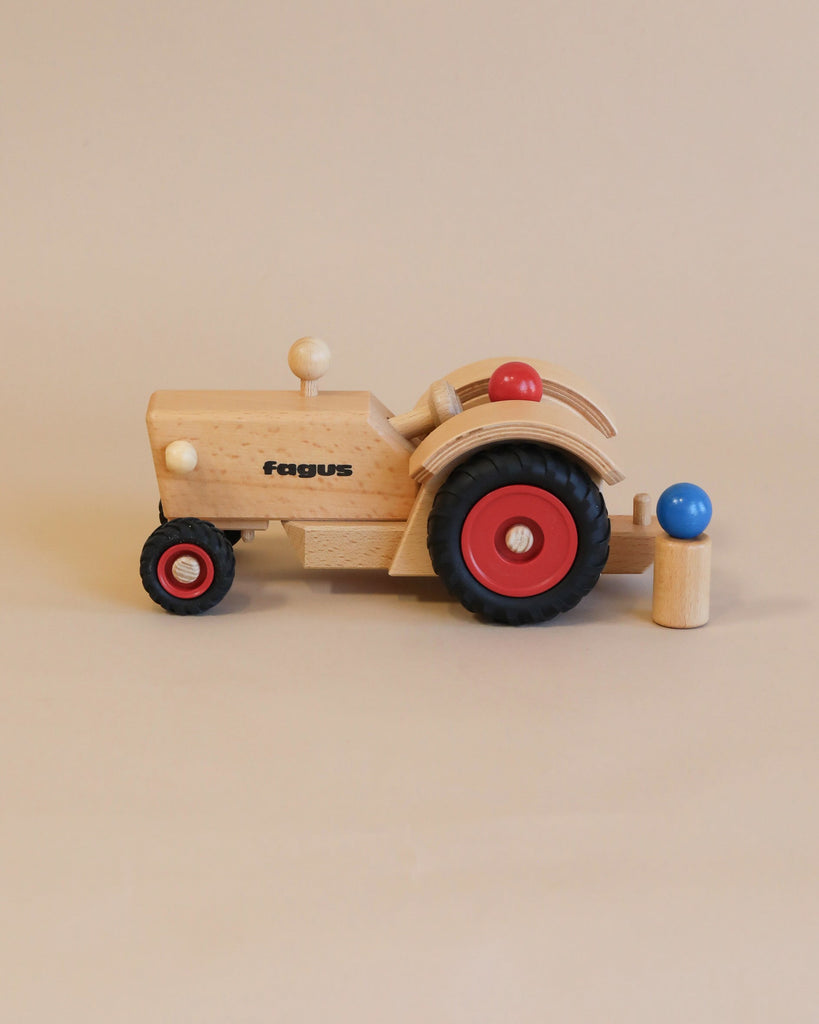 The Maileg x Fagus Tractor Set, featuring handcrafted quality and black and red wheels with the "fagus" logo, stands on a beige background beside a small wooden figure.