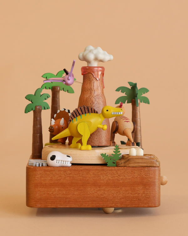 The Wooden Volcano & Dinosaur Music Box features colorful toy dinosaurs, palm trees, a smoking volcano, dinosaur eggs, and a skull on a plain beige background.