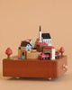 The Wooden Train Music Box - European Town features a miniature village with colorful houses, church, trees, a bridge over water, and a red toy train circling the beige base.