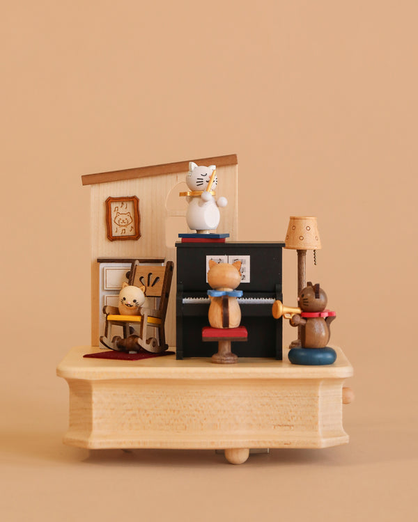 The Wooden Cat Playing Piano Music Box showcases four cat figurines: one at a black piano, one conducting, one in a rocking chair, and one listening, set against a backdrop with a lamp, framed art, and a beige wall.