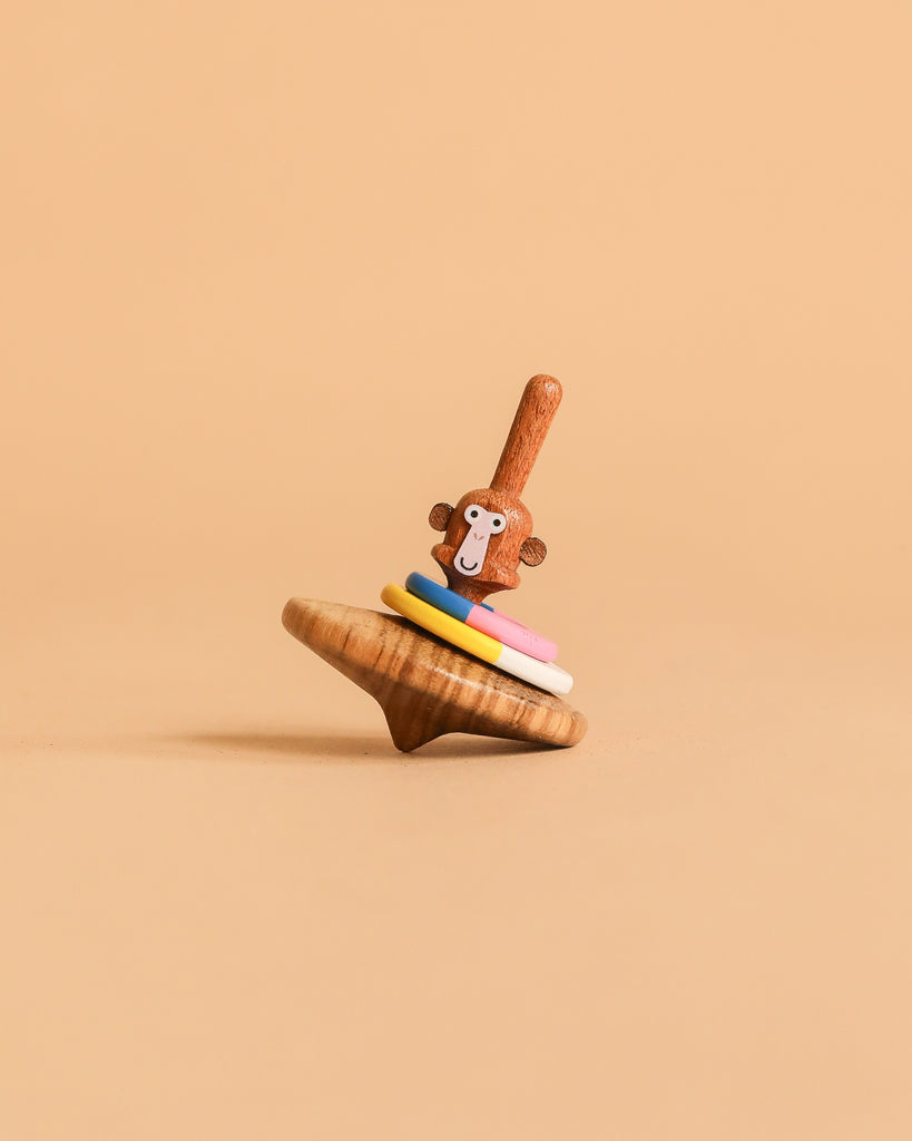 The Wooden Spinning Top - Monkey features a monkey-faced handle and four colorful rings, standing upright against a beige background—an eco-friendly wooden toy providing timeless fun for all ages.