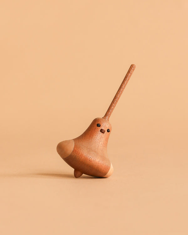 The Wooden Spinning Top - Bird is a small, sustainably sourced wooden top with a cute painted face and long handle, shown standing on a beige surface against a matching beige background.