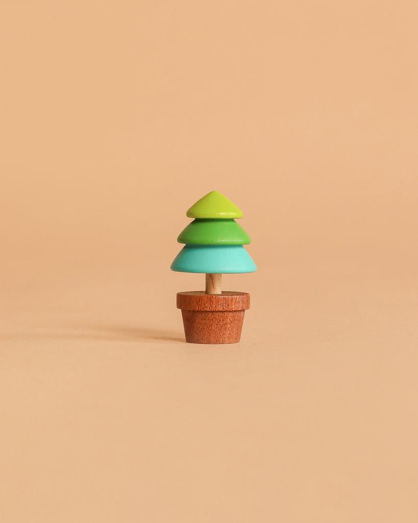 The Wooden Spinning Top - Tree is a small, minimalist wood toy featuring layered green and teal sections, set in a brown pot and shown against a solid beige background.