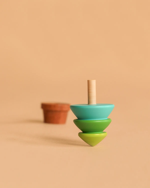 The Wooden Spinning Top - Tree, featuring blue, green, and yellow sections, stands upright on a beige surface with a blurred small brown flowerpot in the background, evoking timeless play.