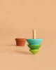 The Wooden Spinning Top - Tree, featuring blue, green, and yellow sections, stands upright on a beige surface with a blurred small brown flowerpot in the background, evoking timeless play.