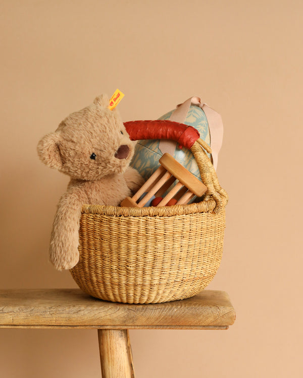 The Easter Basket Set, crafted from elephant grass, contains a plush teddy bear, a floral-patterned light blue pillow, and a wooden toy mallet. It sits on a rustic wooden bench against a beige backdrop.