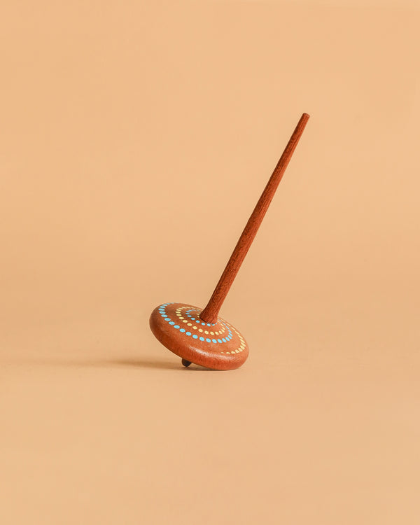 The Wooden Spinning Top - Long Stem, Dots features a sustainably sourced wooden build with white, blue, and yellow dot patterns, standing upright on a beige surface—a classic toy for all ages.
