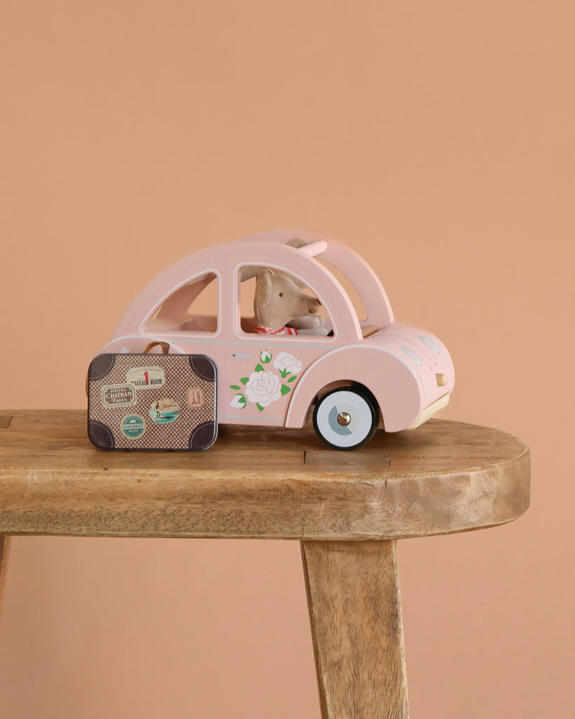 The Maileg Road Trip Adventure Set features a small wooden toy car with a floral design, a plush mouse passenger, and a Maileg suitcase on a stool—perfect for any dolls house scene against a peach background.