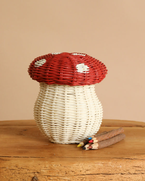 The Olli Ella Mushroom Basket With Coloring Pencils- Red, designed to resemble a red and white mushroom, is placed on a wooden surface with three twig coloring pencils beside it against a solid beige background.